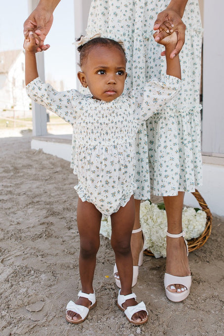 A young girl wears the Ivy City Co Baby Madeline Romper in Blue as she holds an adults hands for support. The adult, in a matching floral dress, stands behind her on sandy ground with a basket of white flowers nearby.