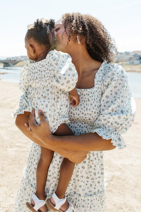 A woman kisses a young girl on the forehead while holding her. Both wear matching Ivy City Co Baby Madeline Rompers in blue, enjoying a sunny day outdoors near water with houses in the background.