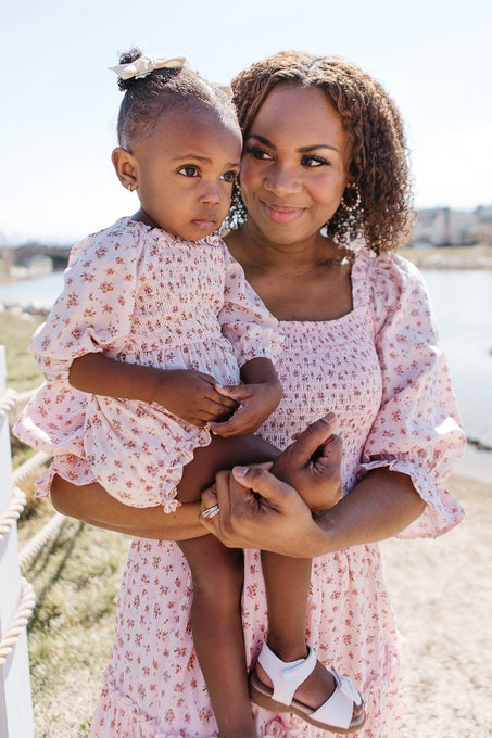 A woman smiles while holding a young girl in the Ivy City Co Baby Madeline Romper in Pink. Both wear matching floral outfits and stand outdoors near water on a sunny day, with the woman gazing at the thoughtful-looking girl.
