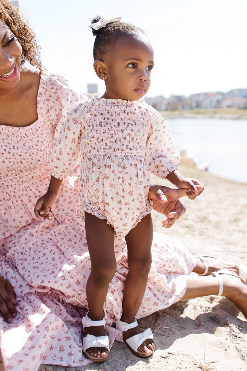 A toddler wears the Ivy City Co Baby Madeline Romper in Pink with white sandals, holding hands with an adult in a matching floral dress by the water on a sunny day.