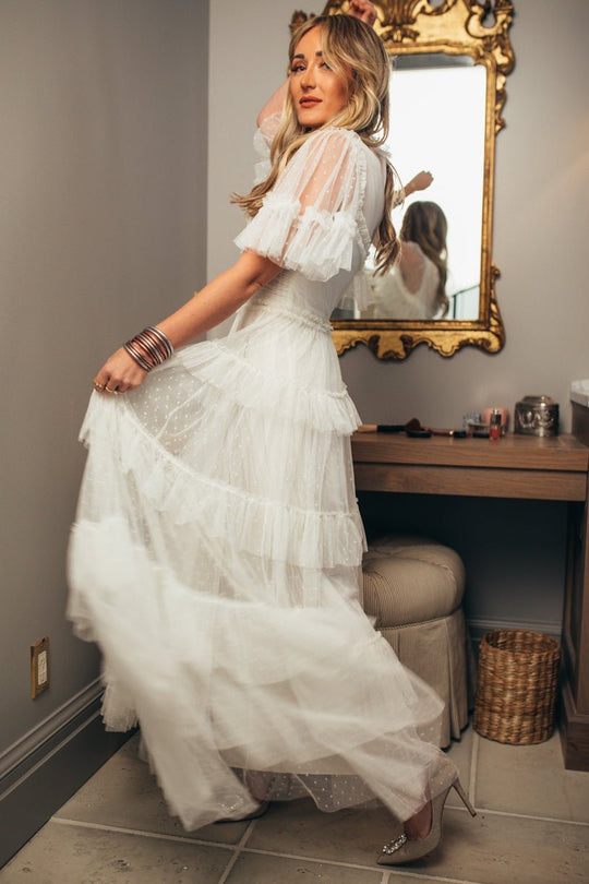 A woman poses indoors by a vanity with a gold mirror, wearing the Ivy City Co. Whimsical Dress in White styled with bracelets and sparkly heels; her blonde hair is in loose waves.