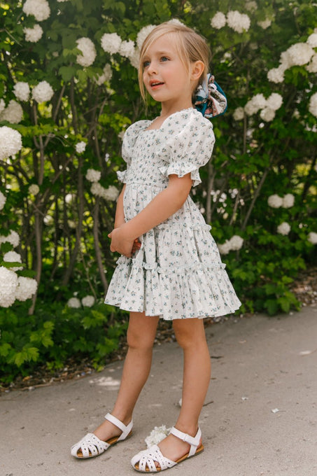 A young girl wears the Ivy City Co Mini Madeline Dress in Blue, featuring a knee-length skirt, puff sleeves, and smocked bodice. She stands on a sidewalk amid blooming white flowers, her blonde hair tied back with a colorful scrunchie.