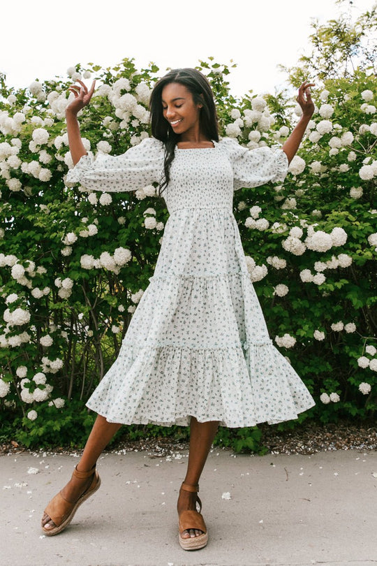 A woman in the Ivy City Co Madeline Dress in Blue dances joyfully outside before blooming white flowers, wearing brown wedge sandals. This maternity-friendly look is perfect for sunny days.