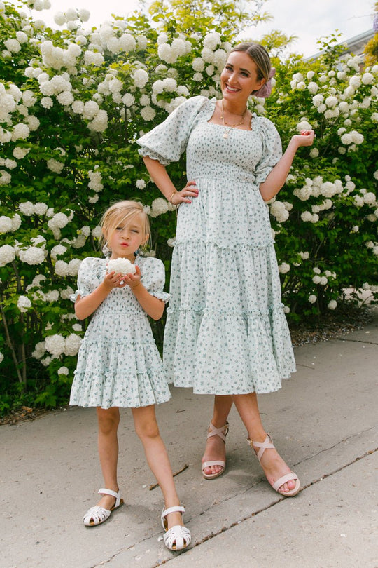 A woman and a young girl smile on the sidewalk in front of blooming white flowers, both wearing matching Ivy City Co Mini Madeline Dresses in Blue with puff sleeves and sandals. The girl holds a flower as they pose for the camera.