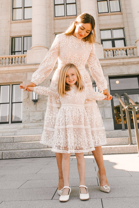 A woman and young girl, both in matching Ivy City Co Mini Sicily Dresses with long sleeves, stand on stone steps and smile, posing together in their elegant white lace midi-length outfits.
