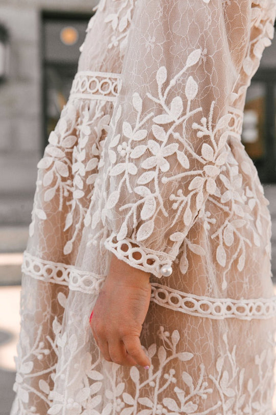 A close-up of a person wearing the Ivy City Co Mini Sicily Dress With Long Sleeve, a white lace midi dress with intricate floral embroidery and circular trim details, standing outdoors with their hand relaxed by their side.