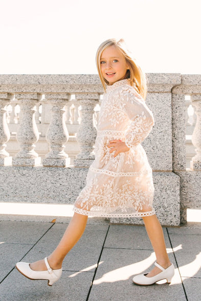 A young girl smiles in bright sunlight while walking on a stone terrace, wearing the Ivy City Co Mini Sicily Dress With Long Sleeve in white lace with champagne lining and matching white shoes.