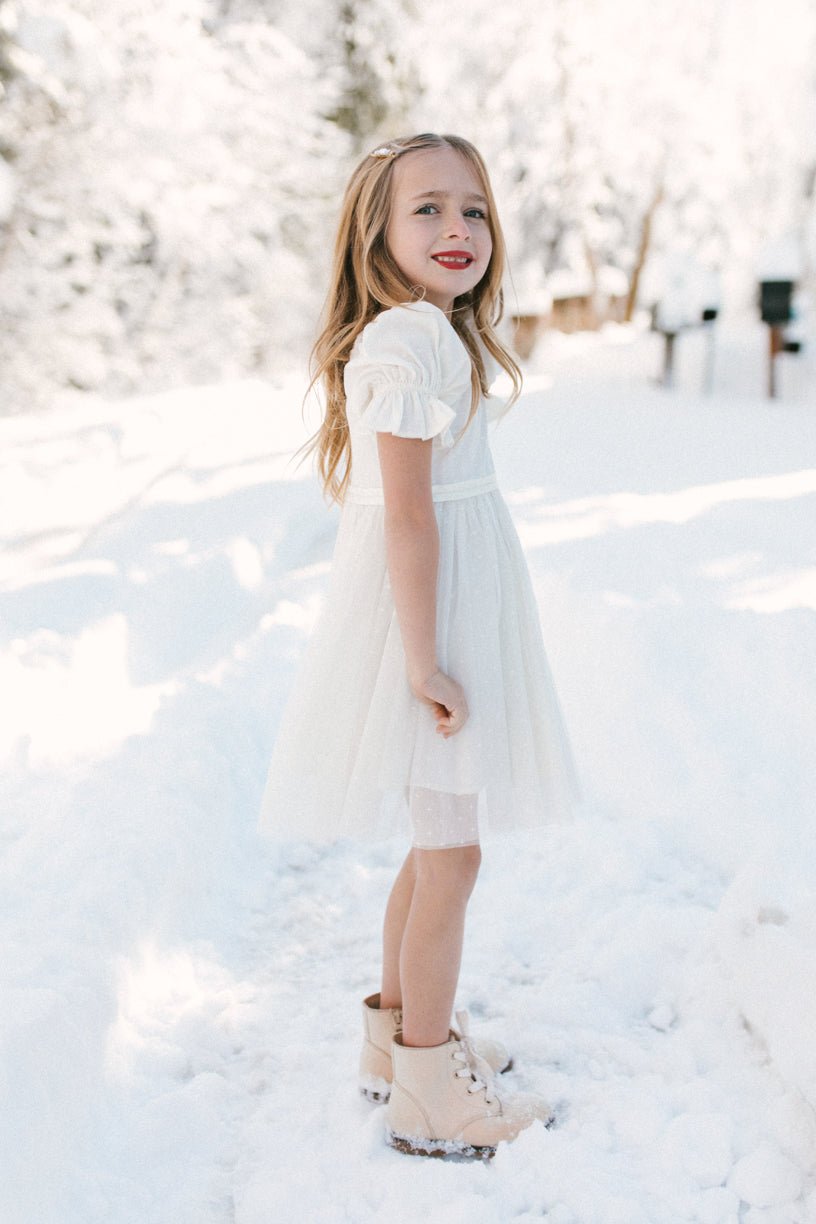 A young girl smiles in the snow, wearing Ivy City Co’s Mini Ballerina Dress in White and beige boots. Her long, wavy blonde hair and the snowy, tree-filled background create a bright winter scene.