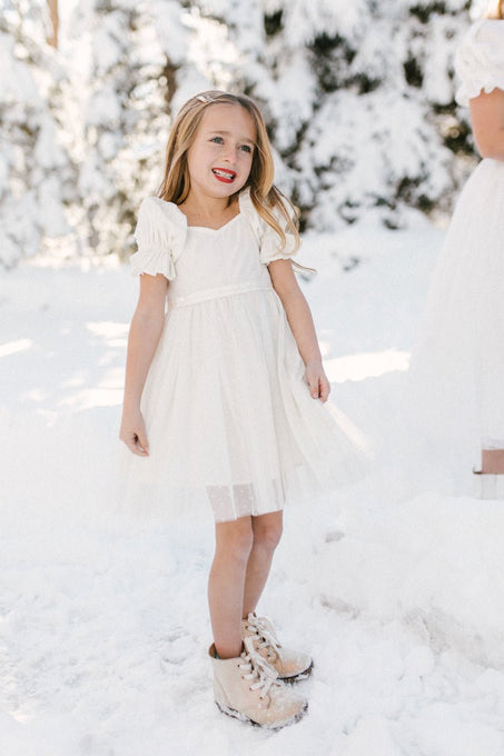 A young girl smiles in the snow wearing the Ivy City Co. Mini Ballerina Dress in White and tan boots, with snowy trees and another person in a white dress visible in the background.