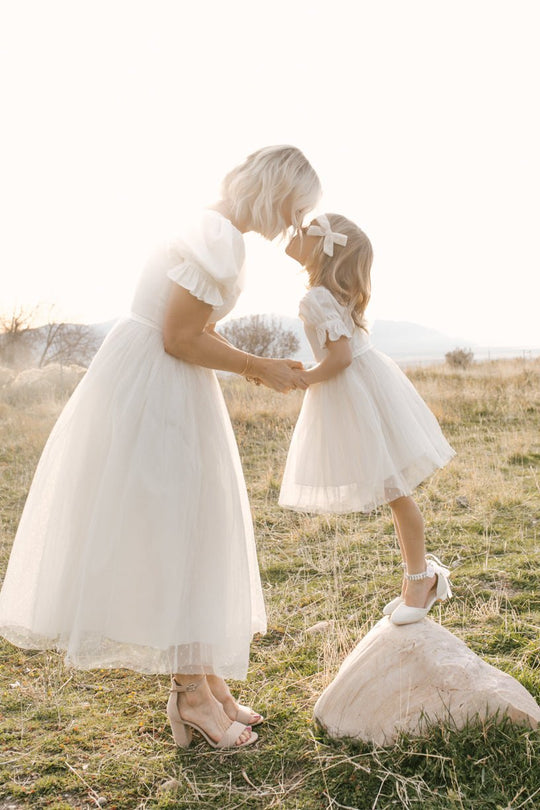 A woman and young girl wear matching Ivy City Co Mini Ballerina Dresses in white, holding hands and touching foreheads on a sunlit field. The girl stands on a rock, capturing a warm and tender outdoor moment.