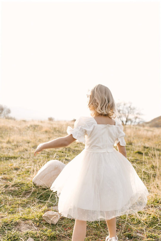 A young girl wearing the Ivy City Co. Mini Ballerina Dress in White stands in a sunlit grassy field, her back to the camera as she reaches toward a rock. The bright, blurred background features sparse trees in the distance.