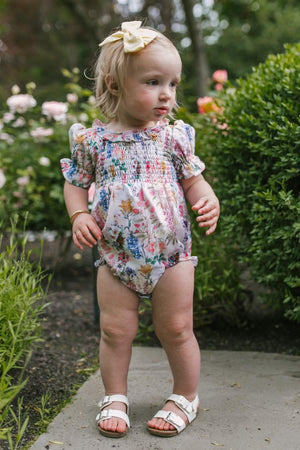 A young toddler with blonde hair, wearing the Ivy City Co Baby Cordelia Floral Romper, white sandals, and a yellow bow, stands on a garden path surrounded by green bushes and blooming flowers.