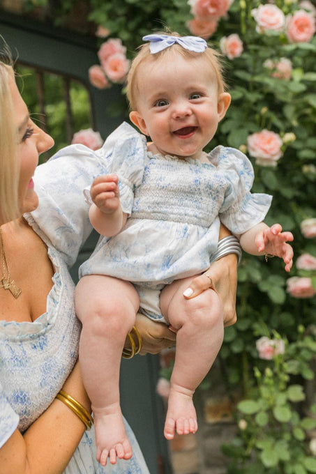 A smiling baby wearing the Ivy City Co Baby Valencia Romper is held by a woman in a matching dress, both standing before a pink rose bush.