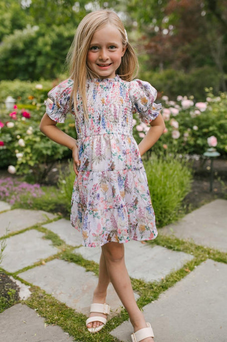 A young blonde girl wearing the Ivy City Co Mini Cordelia Floral Dress and sandals stands on a stone garden path, smiling with her hands on her hips as colorful flowers and lush greenery fill the background.