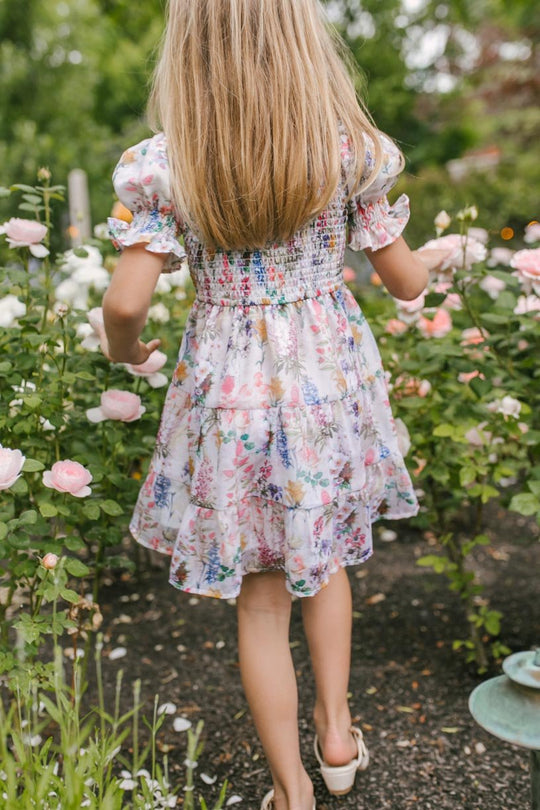A young girl with long blonde hair wears the Mini Cordelia Floral Dress by Ivy City Co as she strolls through a sunny garden filled with blooming pink and white roses.
