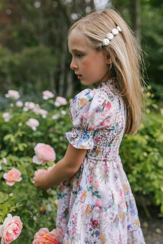 A young girl with long blonde hair and pearl clips wears the Ivy City Co Mini Cordelia Floral Dress as she stands thoughtfully among blooming pink roses in a garden.