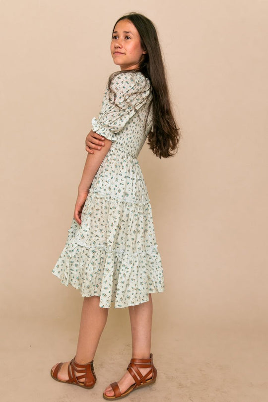 A girl with long brown hair stands sideways, wearing the Ivy City Co Mini Madeline Dress in Blue with puff sleeves and a ruffled hem, paired with brown sandals, posing against a plain beige background.