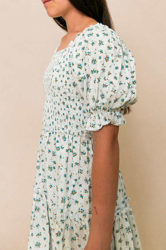 A woman models the Ivy City Co Mini Madeline Dress in Blue, a light blue, knee-length piece with puff sleeves and a smocked bodice, shown in side profile from shoulders to knees against a neutral background.