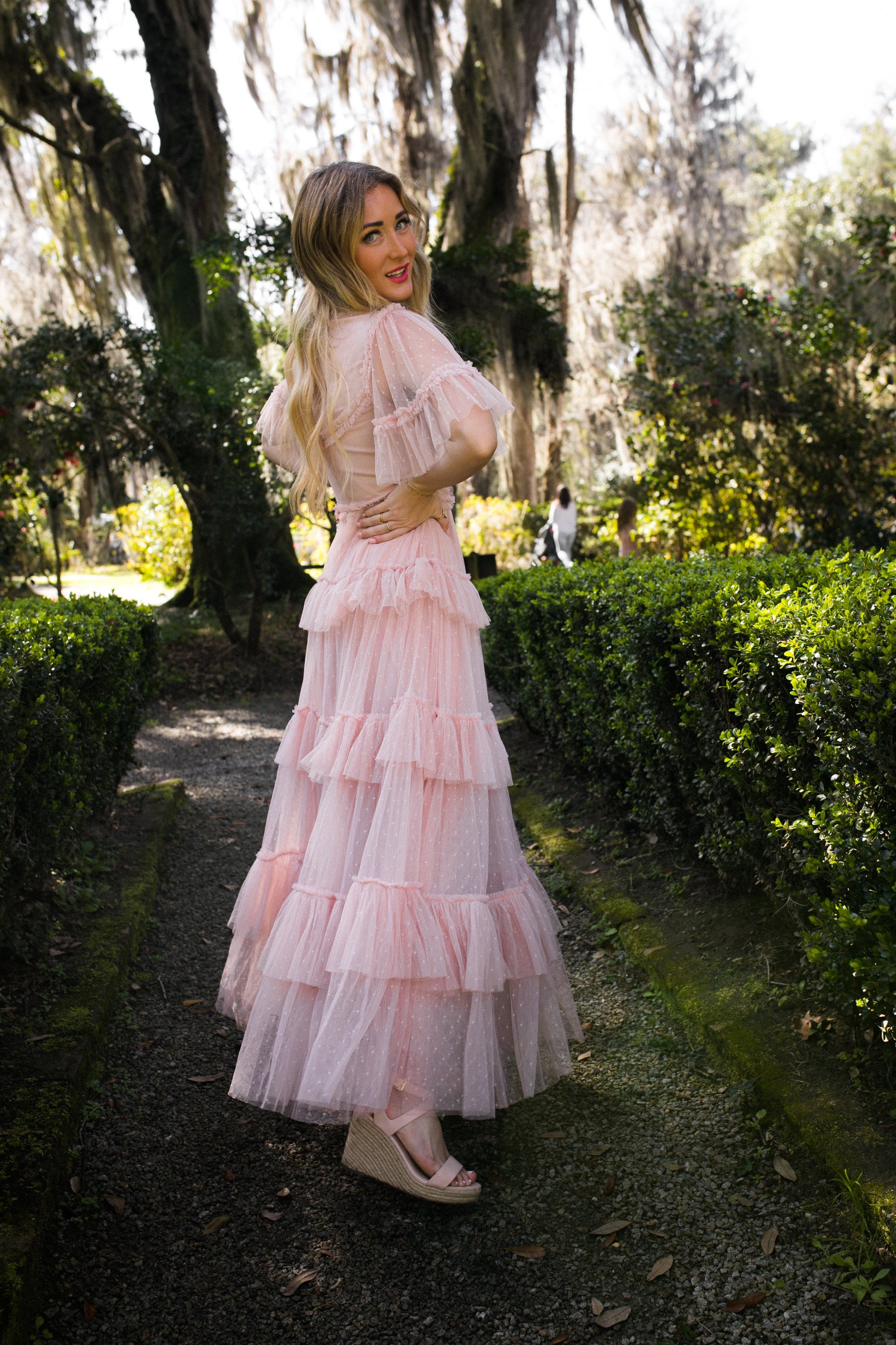 A woman wears the Whimsical Dress in Blush by Ivy City Co, paired with wedge sandals, as she stands on a garden path surrounded by lush greenery and trees in a vibrant outdoor setting.