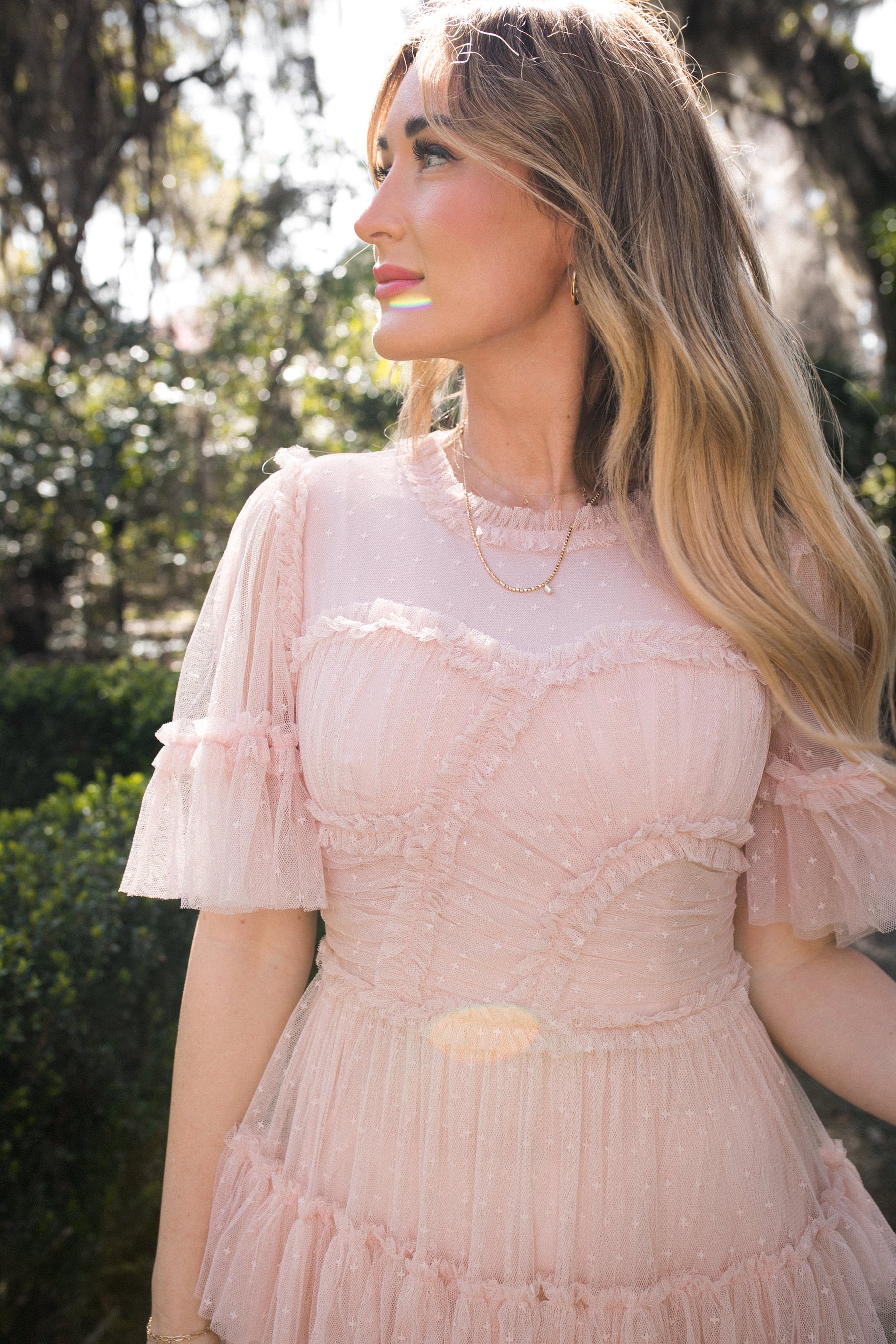 A woman with long blonde hair stands outdoors in sunlight, wearing the Ivy City Co Whimsical Dress in Blush with short sleeves and ruffles. Trees and greenery are blurred in the background.