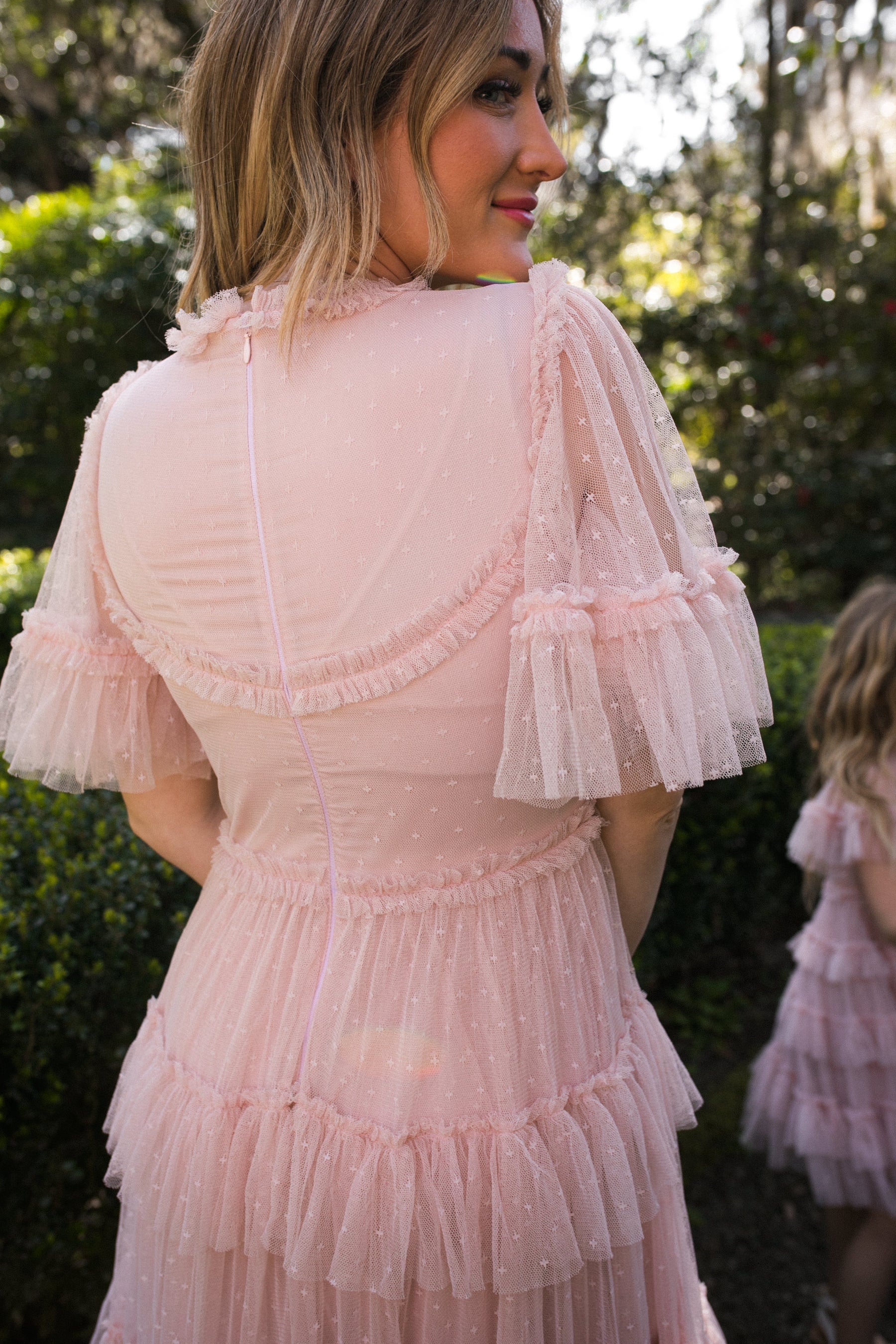 A woman in the Whimsical Dress in Blush by Ivy City Co stands outdoors with her back to the camera, while another girl in a matching dress is seen among greenery and sunlight.