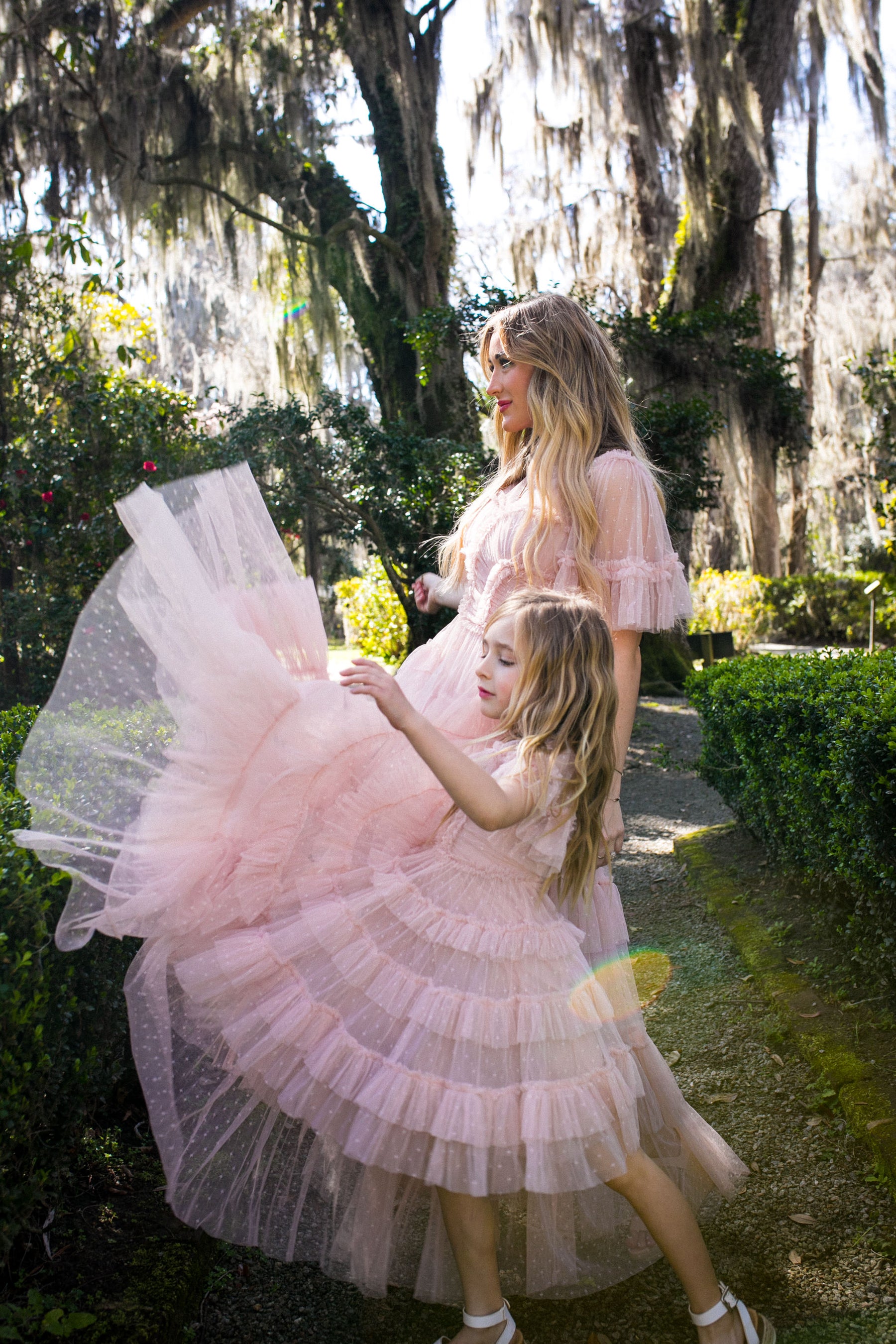 A woman and a young girl wear matching Ivy City Co Whimsical Dress in Blush, strolling along a garden path surrounded by greenery as the girl playfully lifts her dress under the filtered sunlight.