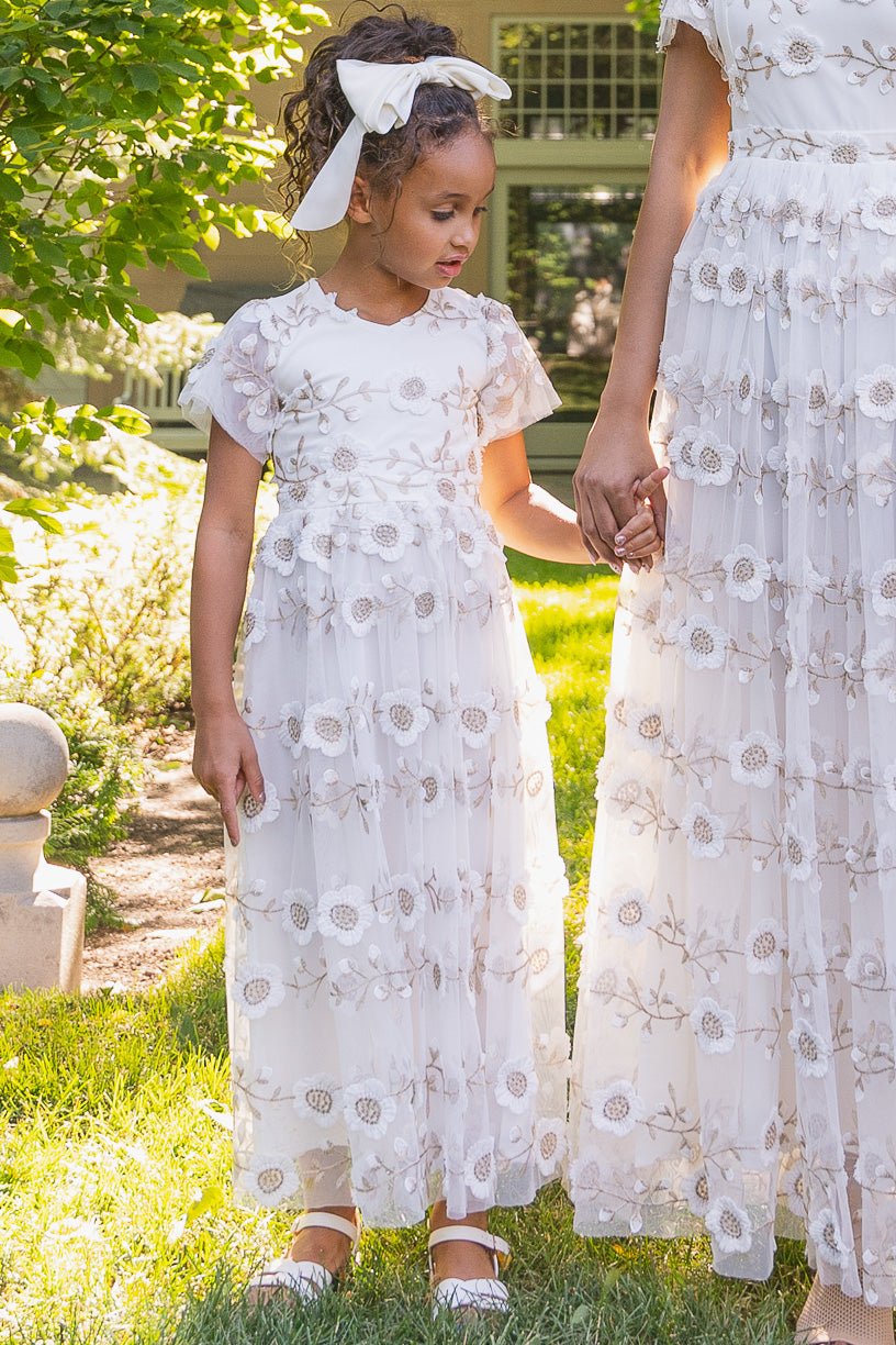 A young girl wears the Ivy City Co Mini Athena Dress in White, holding hands with her mom in matching embroidered outfits, standing together on sunlit grass. The girl's curly hair is tied with a white ribbon and she wears white shoes.