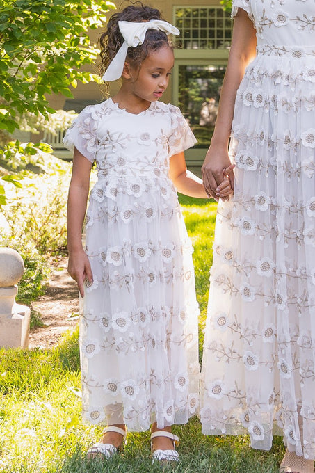 A young girl in the Ivy City Co Mini Athena Dress and white shoes holds hands with an adult in a matching dress, both standing outdoors in a lush, green garden setting.