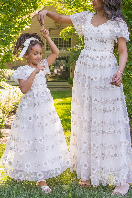 A woman and a young girl wear matching Mini Athena Dresses in White by Ivy City Co as they dance together outside in a sunlit garden, smiling and holding hands.