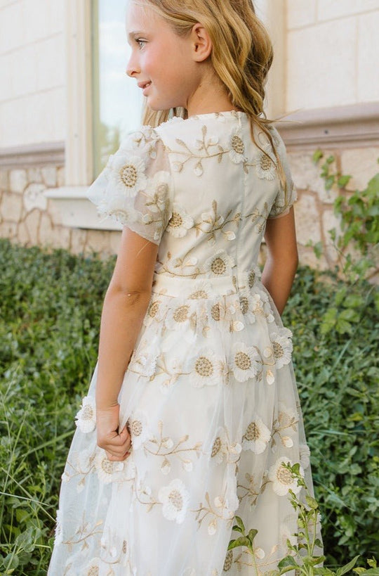 A young blonde girl wearing the Ivy City Co Mini Athena Dress, a white embroidered maxi, stands by green plants and a stone wall outdoors, smiling and looking to the side.