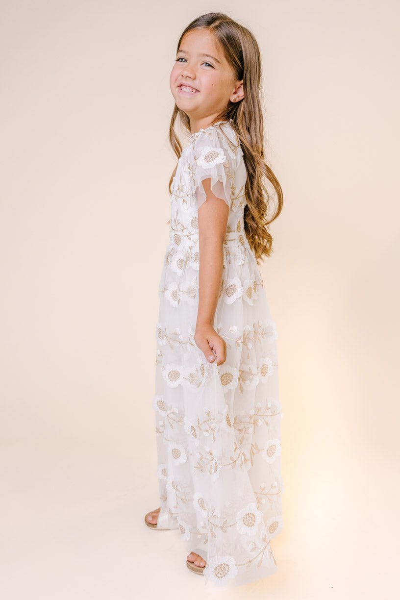 A young girl with long brown hair smiles, standing sideways in the Ivy City Co Mini Athena Dress in White—an ankle-length embroidered dress with floral patterns and short sleeves—against a light beige background.