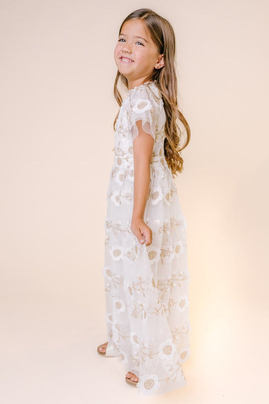 A young girl with long brown hair smiles while wearing the Ivy City Co Mini Athena Dress—a white, floral print maxi with sheer layers and short sleeves—standing sideways against a light beige background.