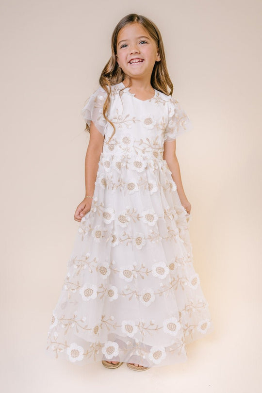A young girl with long brown hair smiles in the Ivy City Co Mini Athena Dress— a maxi-length, short-sleeved white embroidered dress with floral details—standing against a plain beige background.