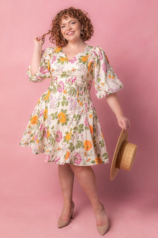 A smiling woman with curly hair poses against a pink backdrop, wearing the Ivy City Co Leah Dress in Multicolor Floral with puffed sleeves, holding a straw hat and standing confidently in nude heels.