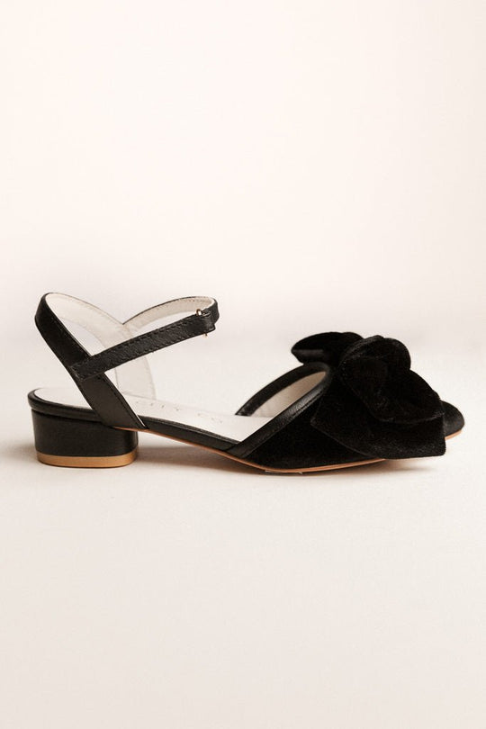 The Ivy City Co Mini Eloise Black Bow Heel is a girls’ closed-toe, low-heeled sandal with an ankle strap and a large decorative black bow on the toe, shown against a plain, light background.