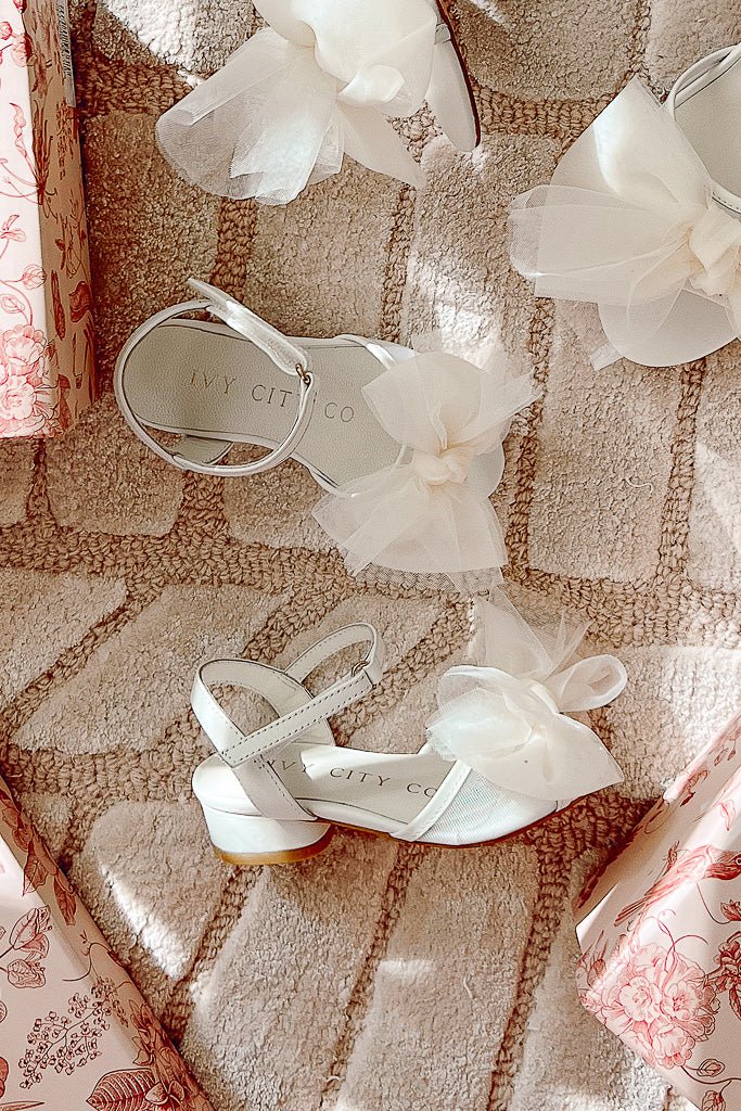 The Mini Ivy Bow Heel in White by Ivy City Co is displayed on a textured beige rug with pink floral gift boxes. These stylish, comfortable sandals for girls feature a large tulle bow detail.