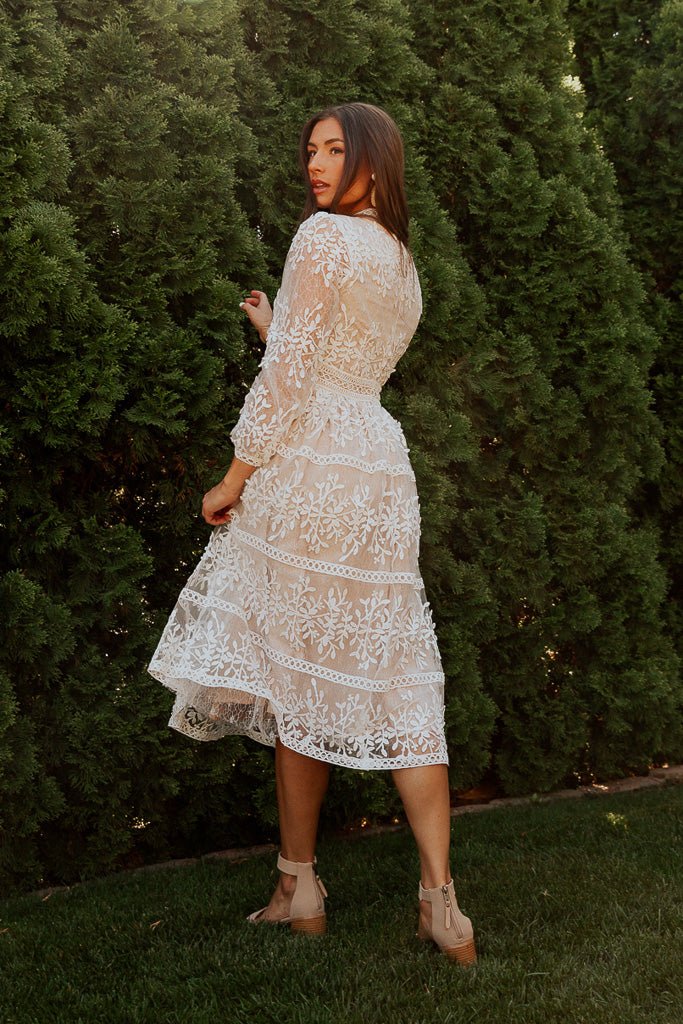 A woman stands on grass in front of tall green bushes, wearing the Ivy City Co Sicily Dress in Champagne Lining—a white lace midi dress with a floral overlay—paired with nude heels, looking back over her shoulder.