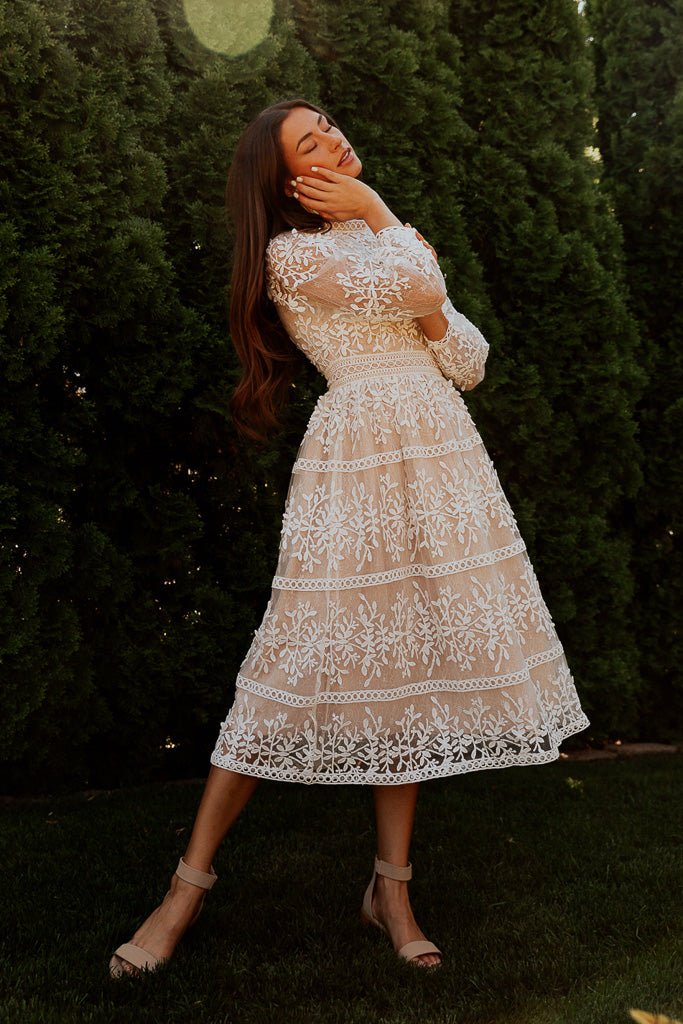 A woman, eyes closed and smiling, stands on grass in the Ivy City Co Sicily Dress in Champagne Lining with a white floral overlay. One hand touches her face, nude heels on her feet, and tall green hedges fill the background.