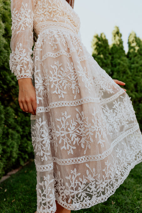 A person stands on grass in the Ivy City Co Sicily Dress - Champagne Lining, a sheer midi dress with an intricate white floral overlay and lace trim. Their face is not visible, and tall green bushes are in the background.