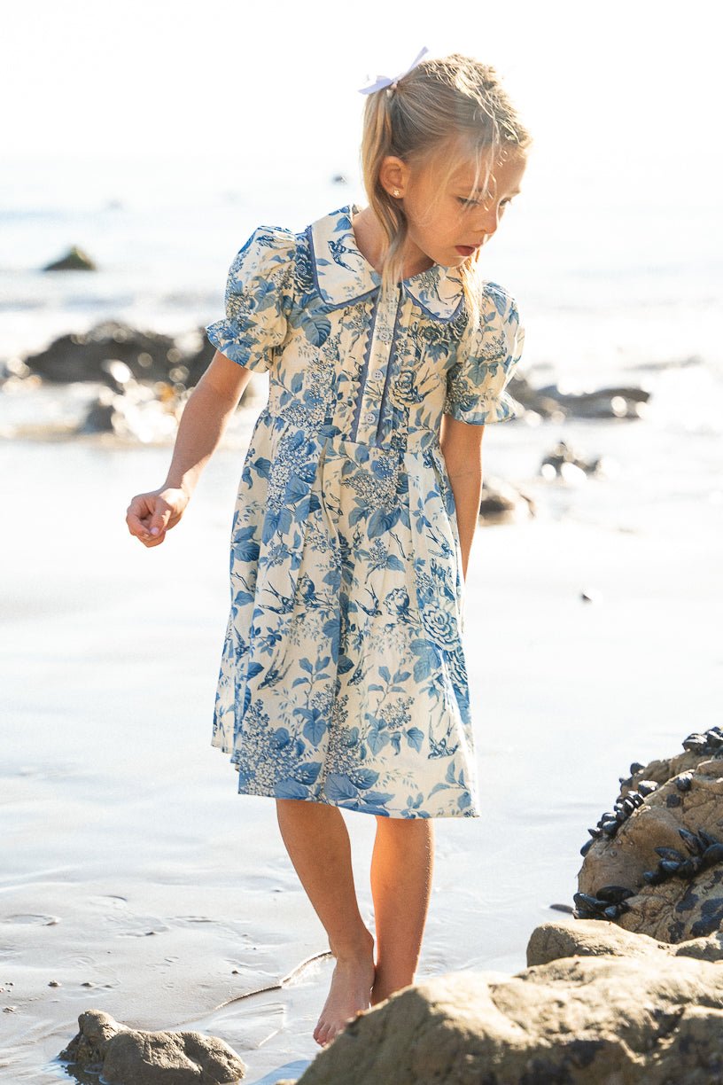 A young girl with blonde hair in a ponytail and a white bow wears the Ivy City Co Mini Sophia Dress in Blue Floral. She walks barefoot near beach rocks, looking down with sunlight behind her.