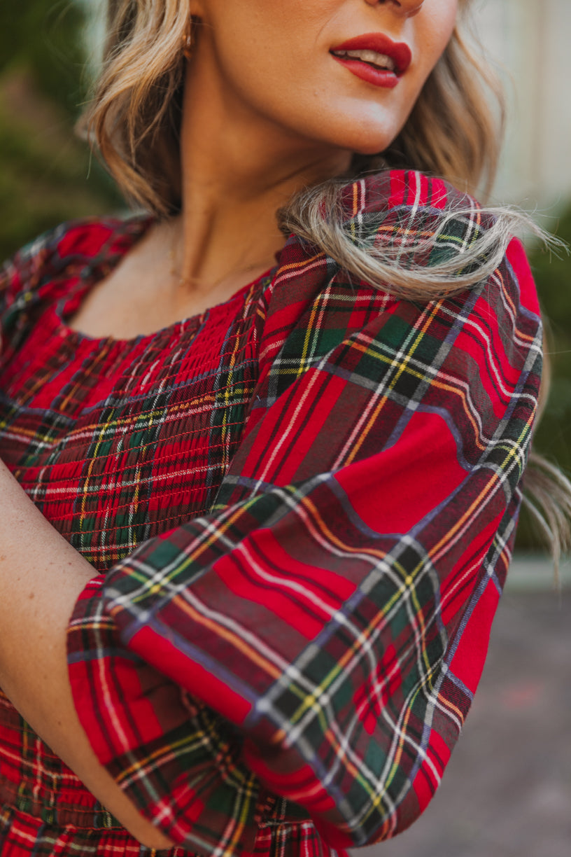A woman stands outdoors wearing the Ivy City Co Madeline Dress in Holiday Plaid, featuring puff sleeves and a maternity-friendly design. Only the lower half of her face and upper body are visible.