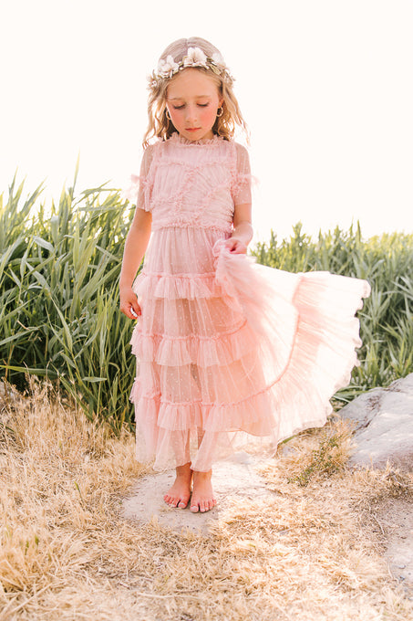 A young girl wears the Ivy City Co Mini Whimsical Maxi Dress in Blush, barefoot on dry grass, lifting one side of her flutter-sleeve tulle skirt. She’s crowned with flowers, surrounded by green plants and bright sunlight.