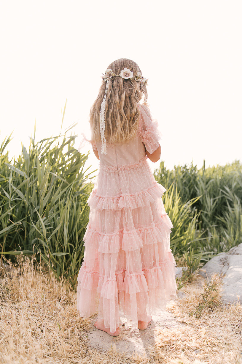 A young girl stands barefoot on dry grass, wearing the Ivy City Co Mini Whimsical Maxi Dress in Blush—long, ruffled with flutter sleeves—and a floral crown in her wavy hair, tall green plants in the background.