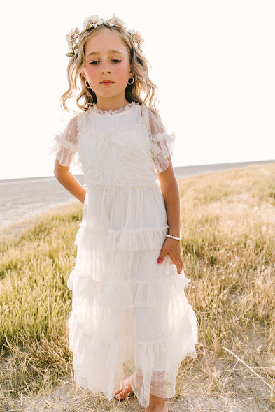 A young girl stands outdoors on dry grass in the Ivy City Co Mini Whimsical Maxi Dress in White, her loose wavy blonde hair adorned with a floral crown. She looks serene and calm, softly illuminated by natural sunlight.