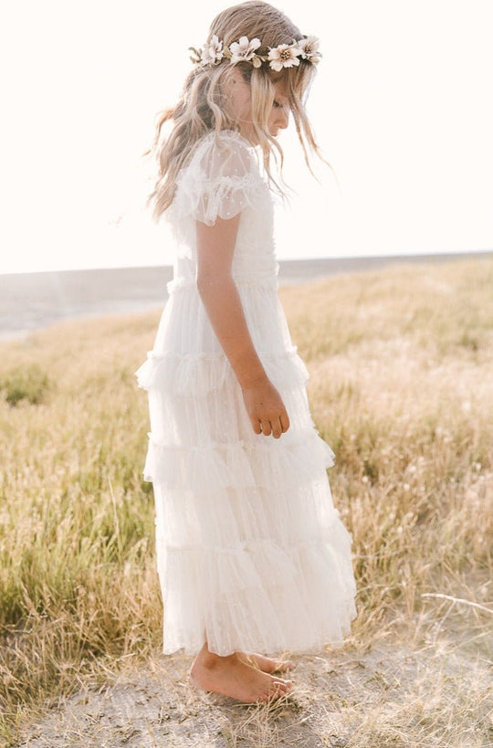 A young girl wears the Ivy City Co Mini Whimsical Maxi Dress in White, featuring flutter sleeves and a layered tulle skirt, as she stands barefoot in a grassy field by the water, sunlight illuminating her flower crown.