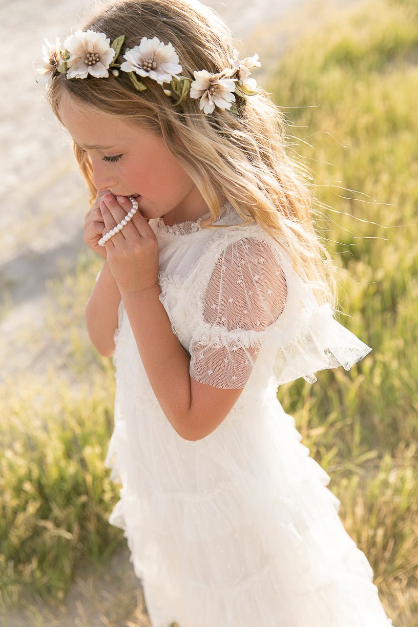 A young girl wears the Ivy City Co Mini Whimsical Maxi Dress in White with flutter sleeves and a flower crown, standing outdoors on green grass and holding a beaded necklace near her face in the sunlight.