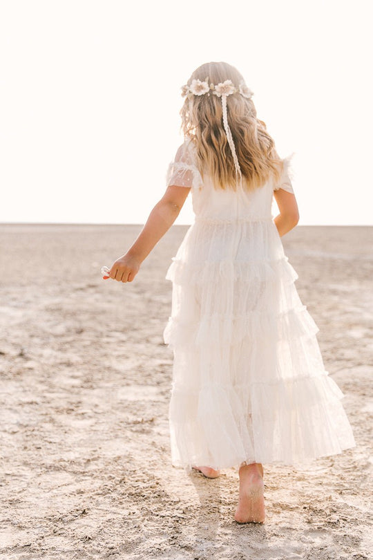 A young girl with long, wavy hair and a floral headband walks barefoot on the beach in the Ivy City Co Mini Whimsical Maxi Dress in White, photographed from behind in soft, natural light.